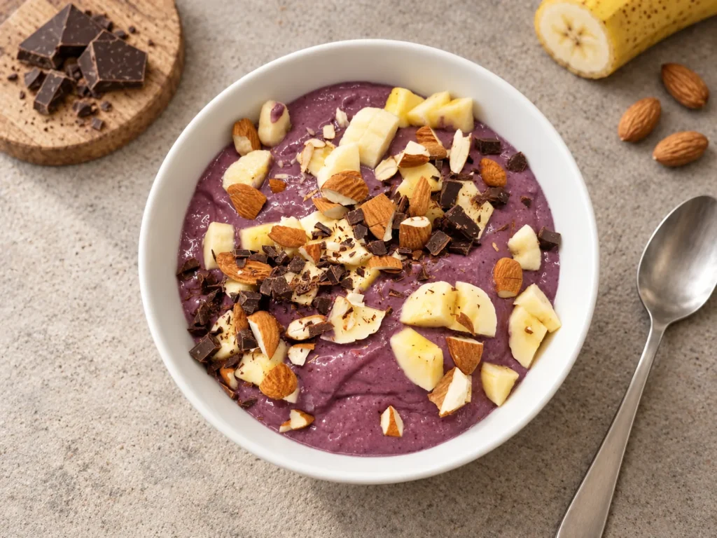 Smoothiebowl mit Banane, Beeren, Nüssen und Schokolade auf einer hellen Tischfläche.